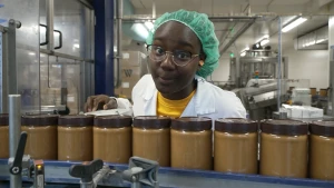 Een foto van presentator Tirsa in een fabriek. Ze heeft een haarnetje op en voor haar langs loopt een lopende band met potjes pindakaas.