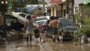 Een foto van 3 mensen op straat in de Filipijnen, auto's liggen op elkaar en de straat staat onder water na de orkaan.
