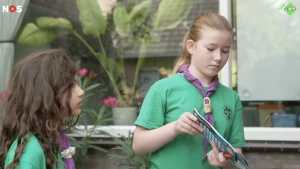 Een foto van twee meisjes in scoutinguniform die een lotenboekje aan iemand buiten beeld laten zien.