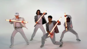 Foto van vier jongens in witte en zilveren kleding die dansen met lichtgevende tl-balken in hun handen.