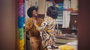 Een foto van een man en een vrouw die dansen in de keuken, ze hebben jaren 70 kleding aan en je ziet slingers hangen.