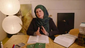 Er is een gesluierde vrouw te zien. Ze zit achter een tafel en heeft dikke boeken voor zich.