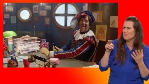 Hoofdpiet kijkt lachend in de camera van achter zijn bureau. Gebarentolk staat naast het scherm.