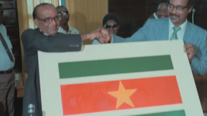 Een foto van twee mannen die elkaar de hand schudden voor zich houden ze een afbeelding van de vlag van Suriname.