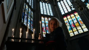 Presentator (vrouw) houdt een lange kaars vast in de kerk. Achter haar zijn glas-in-loodramen zichtbaar.
