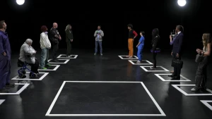 Een foto van een zaal. Op de grond zijn met tape hokjes afgeplakt. Allerlei verschillende mensen staan in de hokjes, zoals een oude vrouw met een rollator, een man in pak en een jonge alternatief geklede vrouw. In het midden is ook een groot leeg hokje.