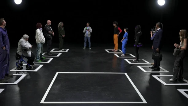 Een foto van een zaal. Op de grond zijn met tape hokjes afgeplakt. Allerlei verschillende mensen staan in de hokjes, zoals een oude vrouw met een rollator, een man in pak en een jonge alternatief geklede vrouw. In het midden is ook een groot leeg hokje.