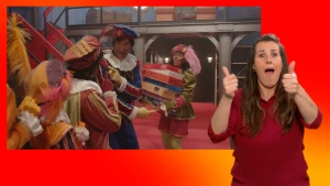 Een Piet staat met een stapel puzzeldozen. Hoofdpiet is in gesprek met andere Pieten. Naast het scherm staat de gebarentolk.