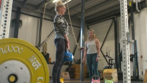 Een foto van presentator Janouk in een sportschool. Ze tilt een kettlebell op. Een trainer staat toe te kijken.