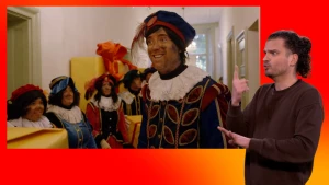 Hoofdpiet staat in de gang van Het Grote Pietenhuis. Om hem heen staan Pieten in een rij. Naast het scherm staat een gebarentolk.
