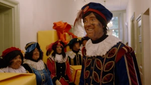 Hoofdpiet staat in de gang van Het Grote Pietenhuis. Om hem heen staan Pieten in een rij.