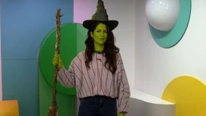 Een afbeelding van de presentatrice met een groen gezicht, een heksenhoed op en een stok in haar hand, naar een personage uit de film Wicked