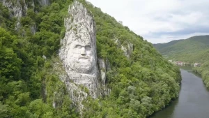 Een foto van een Roemeens landschap met links van de rivier een uit de rotswand uitgehouwen hoofd van een koning van de Daciers.