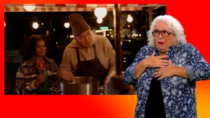 Stephanie staat naast een man die in de soep roert. Naast het scherm staat de gebarentolk.