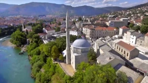 Een foto van een stad in Bosnië en Herzegovina met op de voorgrond een moskee met minaret, links stroomt een rivier en op de achtergrond zie je bergen.