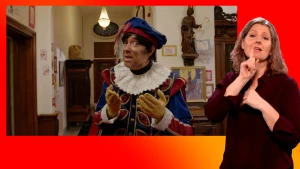 Hoofdpiet gebaart naar de camera. Naast het scherm staat de gebarentolk.