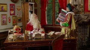 Sinterklaas zit achter zijn bureau. Piet draagt een stapel boeken.