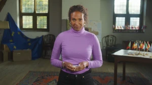 Presentatrice Astrid staat op een zolder. Naast haar een EU vlag en een tafel met vlaggetjes erop.