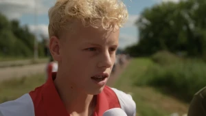 Een foto van een jongen met een Ajax shirt. Hij wordt geïnterviewd en spreekt in een NOS microfoon.