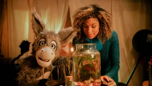 Een foto van de presentatrice en het ezeltje, ze zitten naast elkaar aan tafel, de presentatrice kijkt in een vissenkom die op tafel staat.