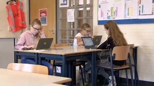 Een afbeelding van drie meisjes in een klaslokaal. Zij werken alle drie op hun laptop en dragen oortjes.