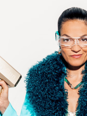 Een foto van de presentatrice met een lichtblauw jasje aan met een grote zwarte bontkraag. In haar hand heeft ze een apparaat en haar andere hand heeft ze in haar zij gezet.