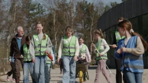 Een afbeelding van een groep kinderen in de natuur. Ze dragen groene hesjes en hebben prikstokken en een kruiwagen mee.