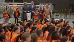 Een afbeelding van het Koningspaar omringd door dansende kinderen in oranje hesjes.