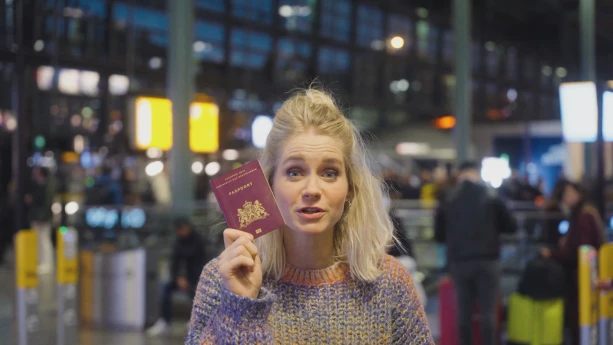 Een foto van presentatrice Janouk met een paspoort in haar hand, ze houd het paspoort naast haar gezicht.