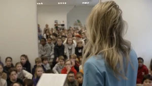 Een afbeelding van een heel groep kinderen zittend op de grond, voor hen staat een mevrouw met een boek in haar hand. Zij leest voor.