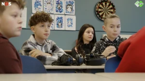 Een foto van 4 leerlingen die aan hun tafeltjes in een klas zitten. Op de tafels liggen koptelefoons.