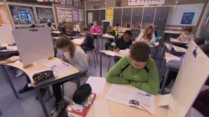 Een foto van een klaslokaal met kinderen die een toets maken, voor hen ligt een boekje op tafel en tussen de tafel staat een schot, zodat ze niet kunnen spieken.