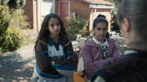 Een foto van twee meisjes op straat, ze staan met hun armen over elkaar, op de rug zien we nog een derde persoon.