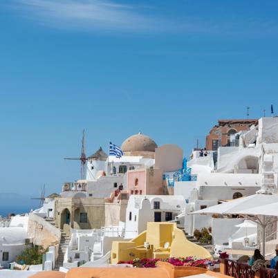 Een foto van een Grieks eiland met witte en gele huisjes tegen een blauwe lucht, ook staan er 2 Griekse vlaggen.