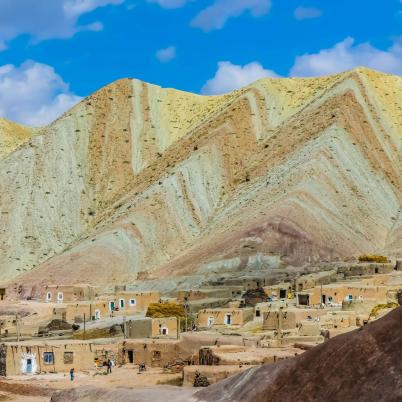 Een foto van een berg of afgraving in Iran, je ziet verschillende gekleurde lagen in de berg, op de voorgrond staan huizen.