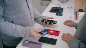 Een foto van twee mensen aan weerszijden van een balie, je zet alleen hun buik en handen, op de balie liggen een aantal mobieltjes. De persoon links houdt een telefoon vast die opengemaakt is en in zijn andere hand de batterij.
