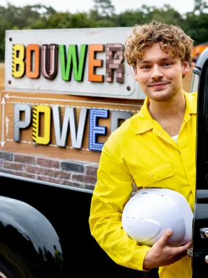 Foto van Soy Kroon die uit een auto stapt. Achterop de auto staat een bord met daarom "Bouwer Power" geschreven.