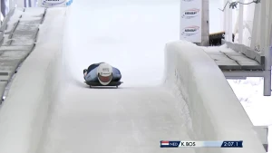 Foto van skeletonatleet die liggend op zijn buik op een slee van een bevroren bobsleebaan suist. Hij draagt een witte helm en een lichtblauw pak.