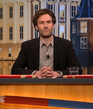 Foto van presentator Roel Maalderink achter de desk van Kiespijn, grote rode potloden liggen voor hem en achter hem een backdrop van het torentje in Den Haag.