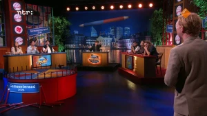 Still uit een aflevering van Kiespijn met de presentatordesk in het midden en de deelnemersdesks links en rechts daarvan. 