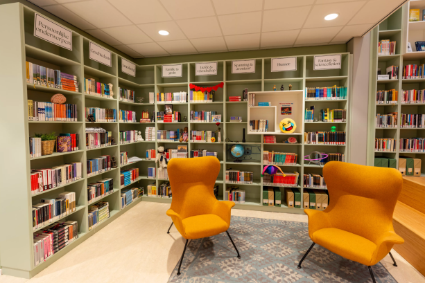 Een foto van een mediatheek in een school met lichtgroene boekenkasten vol boeken en objecten, zoals een wereldbol. Voor de kast staan twee gele stoelen. In de kasten hangen bordjes met genres, zoals 'Familie & gezin', 'Humor' en 'Liefde & vriendschap'.